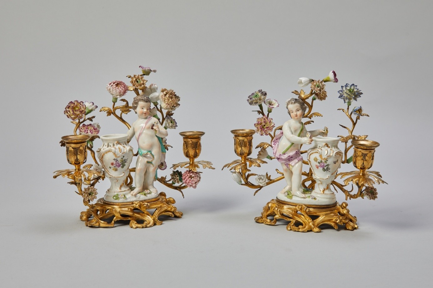 A pair of double-flamed table candlesticks with cherubs. A pair of double-flamed table candlesticks with cherubs<br />
<br />
German, Meissen, circa 1750/1760<br />
<br />
Johann Joachim K&auml;ndler (1706 Fischbach/Arnsdorf &ndash; 1775 Meissen)<br />
<br />
Porcelain, polychrome painting, gold decoration, bronze mounting, Vincennes flowers<br />
<br />
Blue sword mark<br />
<br />
On a curved, slightly raised rocaille base, figure of a cherub standing next to a vase. Behind it, symmetrically extending branches with Vincennes flowers and spouts.&nbsp;<br />
<br />
&nbsp; | Ralph Gierhards Antiques & Fine